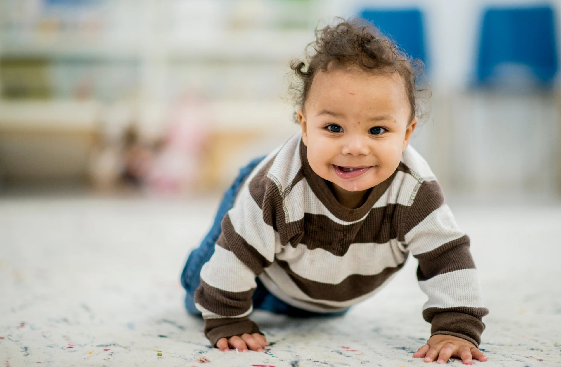 A baby boy crawling and smiling towards the camera. He is wearing a rugby-style shirt with brown stripes.