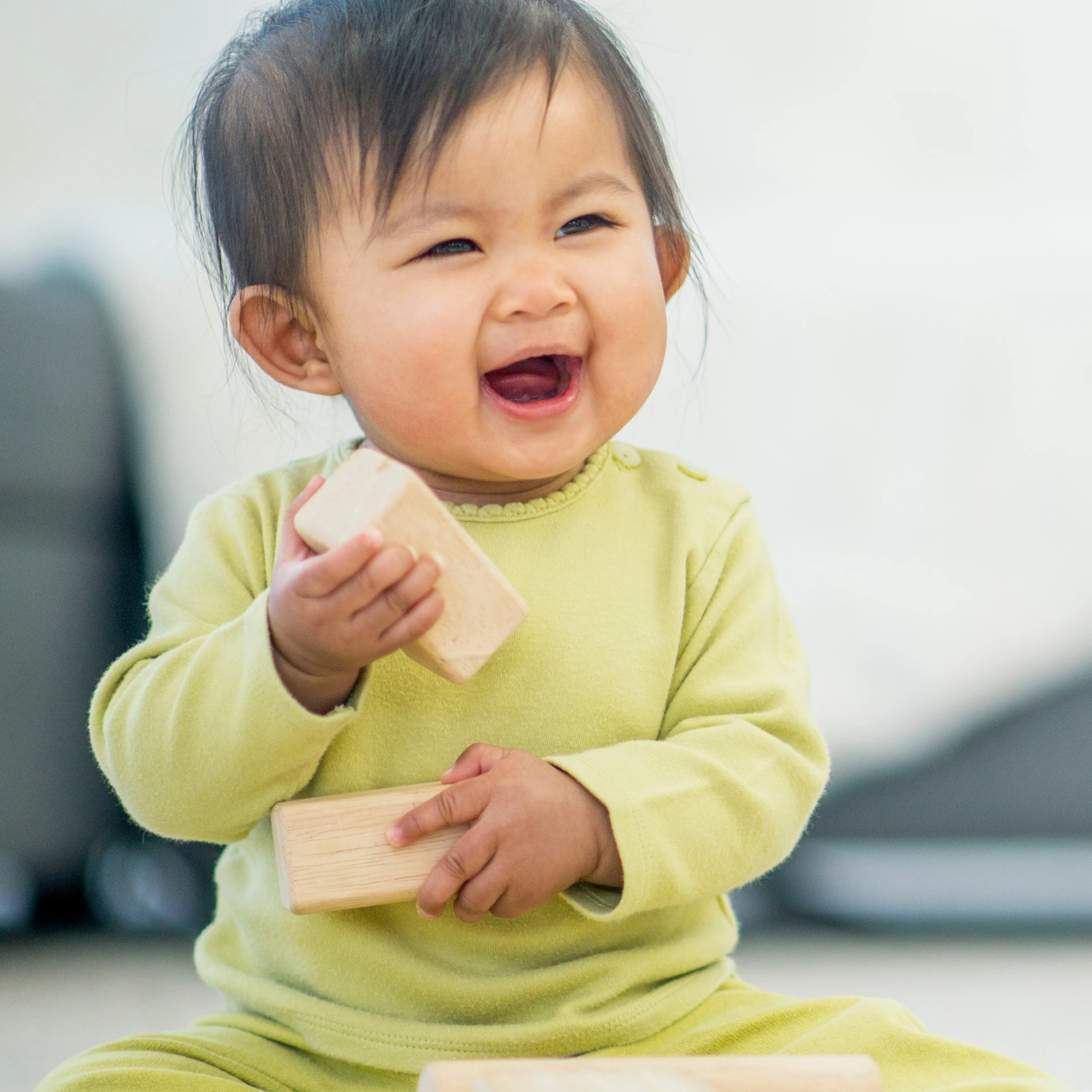 Asian baby in a light green outfit and playing with wood blocks. Baby is smiling with their mouth wide open.