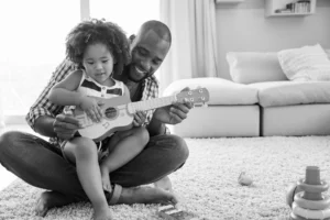 African American father sitting cross-legged on the floor. His preschool daughter is sitting on his lap, and they are playing a guitar together.
