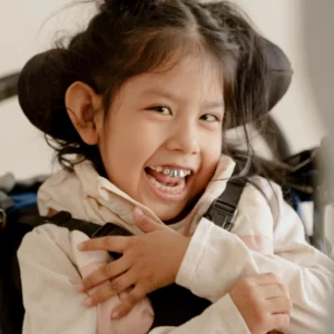 A young girl with light tan skin smiling. She's disabled and uses a wheelchair.