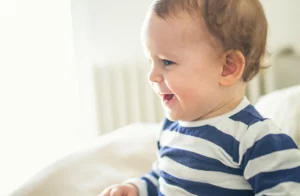 A baby boy smilining to the left of camera. He's wearing a blue and white striped shirt.