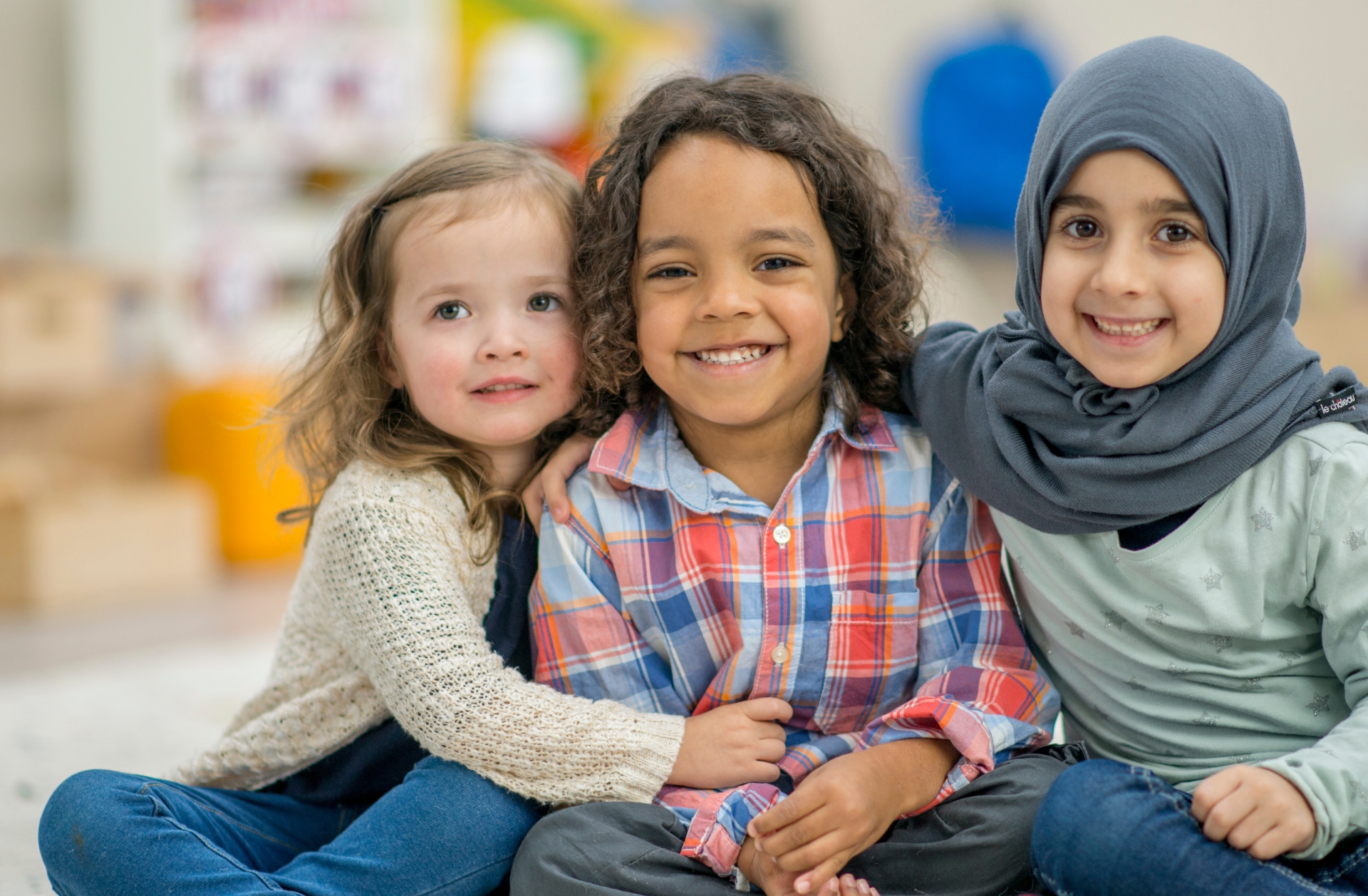 A group of three children (a white girl, a boy with brown skin, and a Muslim girl) sitting together in a preschool.