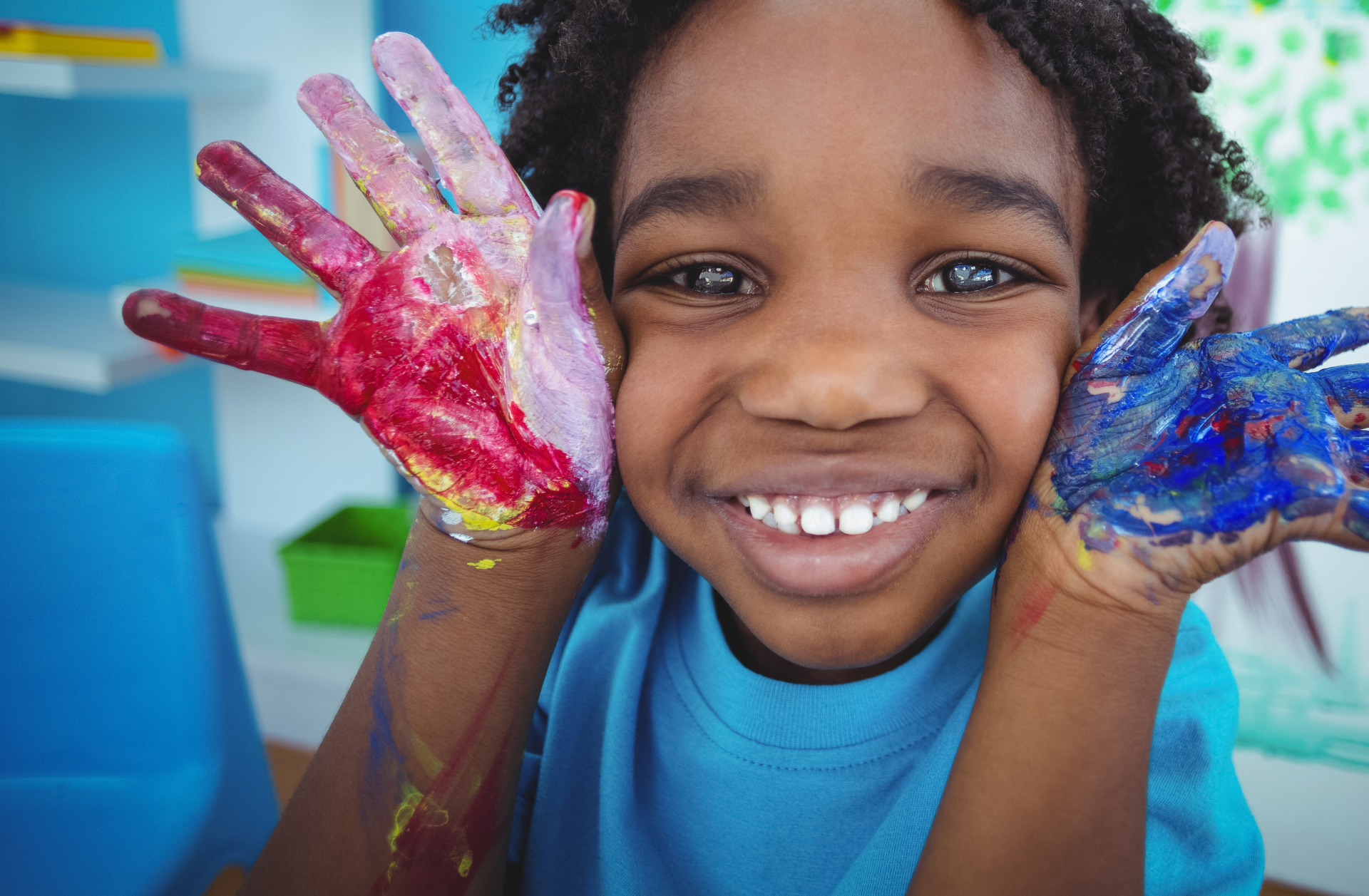 A young Black boy in art class. He's smiling closeup to the camera lens with red paint on his left palm and purple paint on his right palm.