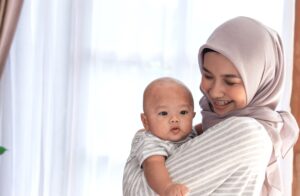 A young mom, wearing a light pink hijab, holding her infant son.