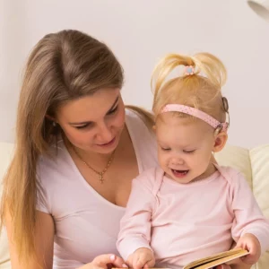A mom reading with her toddler daughter. Both are wearing pink, have fair skin, and the daughter has a cochlear implant visible.