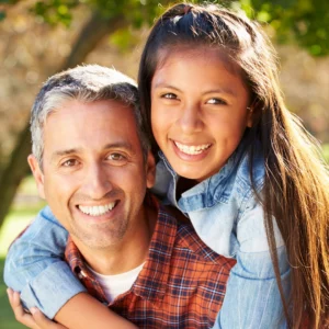 A Latino father with his tween daughter. The daughter has her arms wrapped around her dad from behind and both are smiling at the camera.