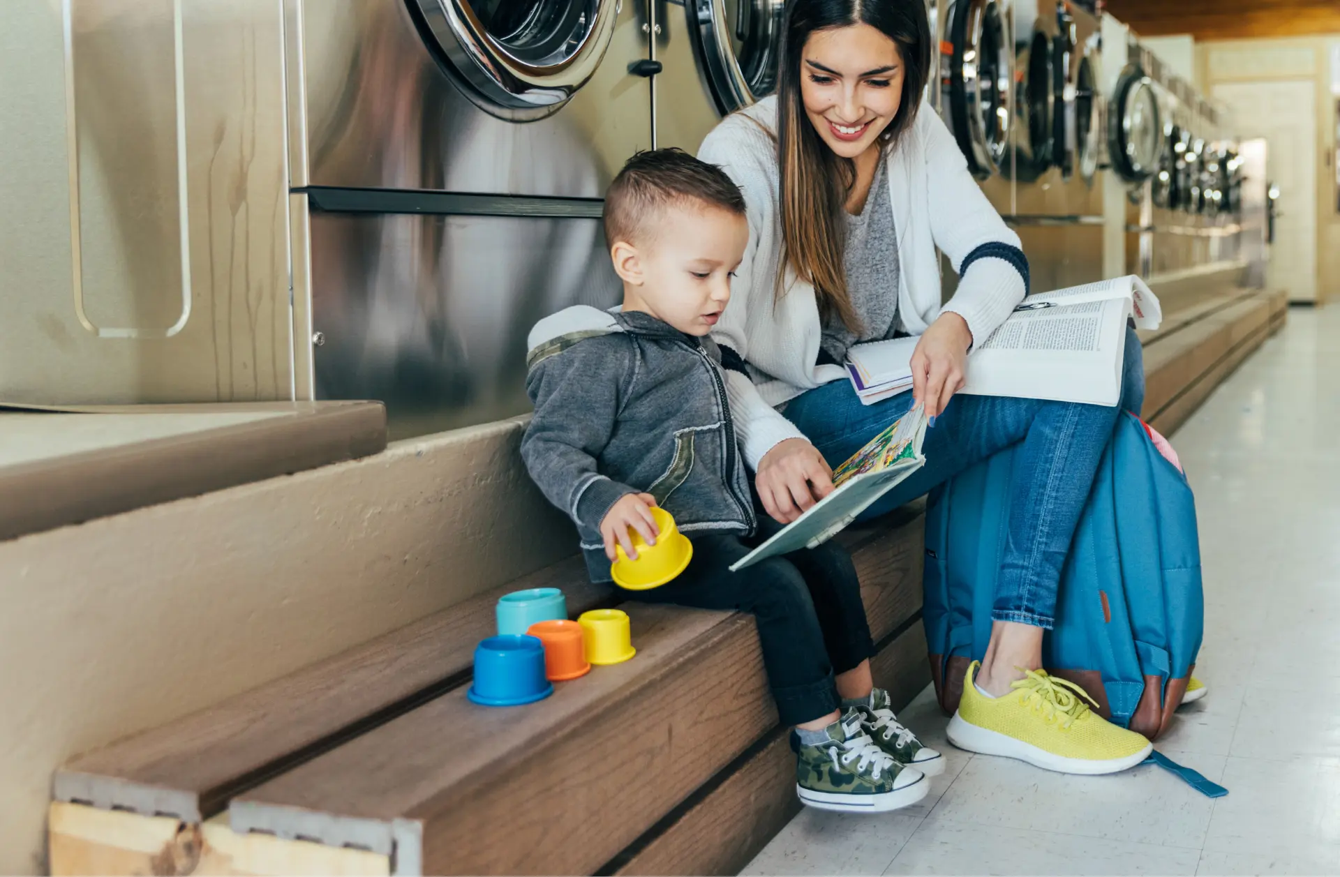 A young mom sitting in front of laundry machines with her toddler son. She has a textbook in her lap, but she's leaned over and reading his book to him. The boy is smiling and holding big colorful blocks.