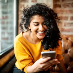 A woman of color smiling at her phone in a coffee shop. She's wearing a mustard yellow sweater and has curly black hair.