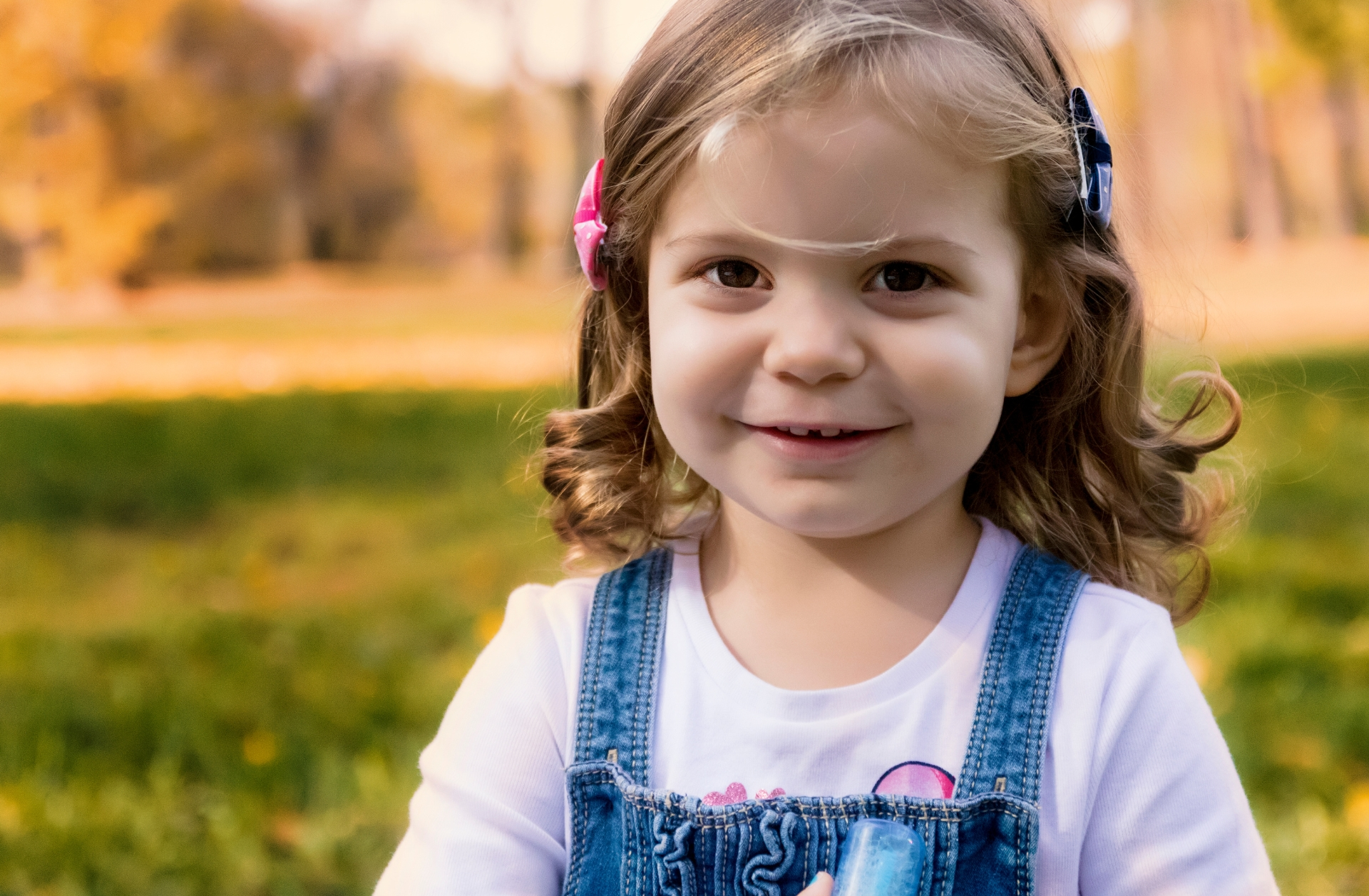 A very young girl smiling and standing outside on grass. She's wearing a denim jumper dress and a pink bow in her hair.