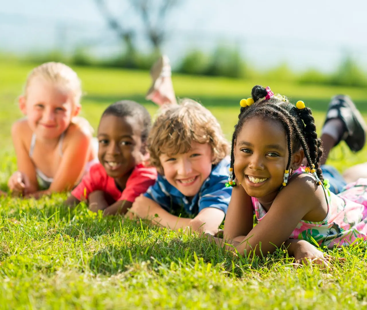 Four children laying on their bellies in the grass. All of them are smiling at the camera. From left to right, the children are white, Black, white, and Black.