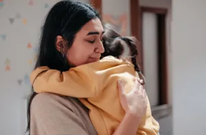 A mother with a peaceful expression hugs her young child, who is wearing a yellow sweatshirt. They share a close, warm embrace, conveying love and support.