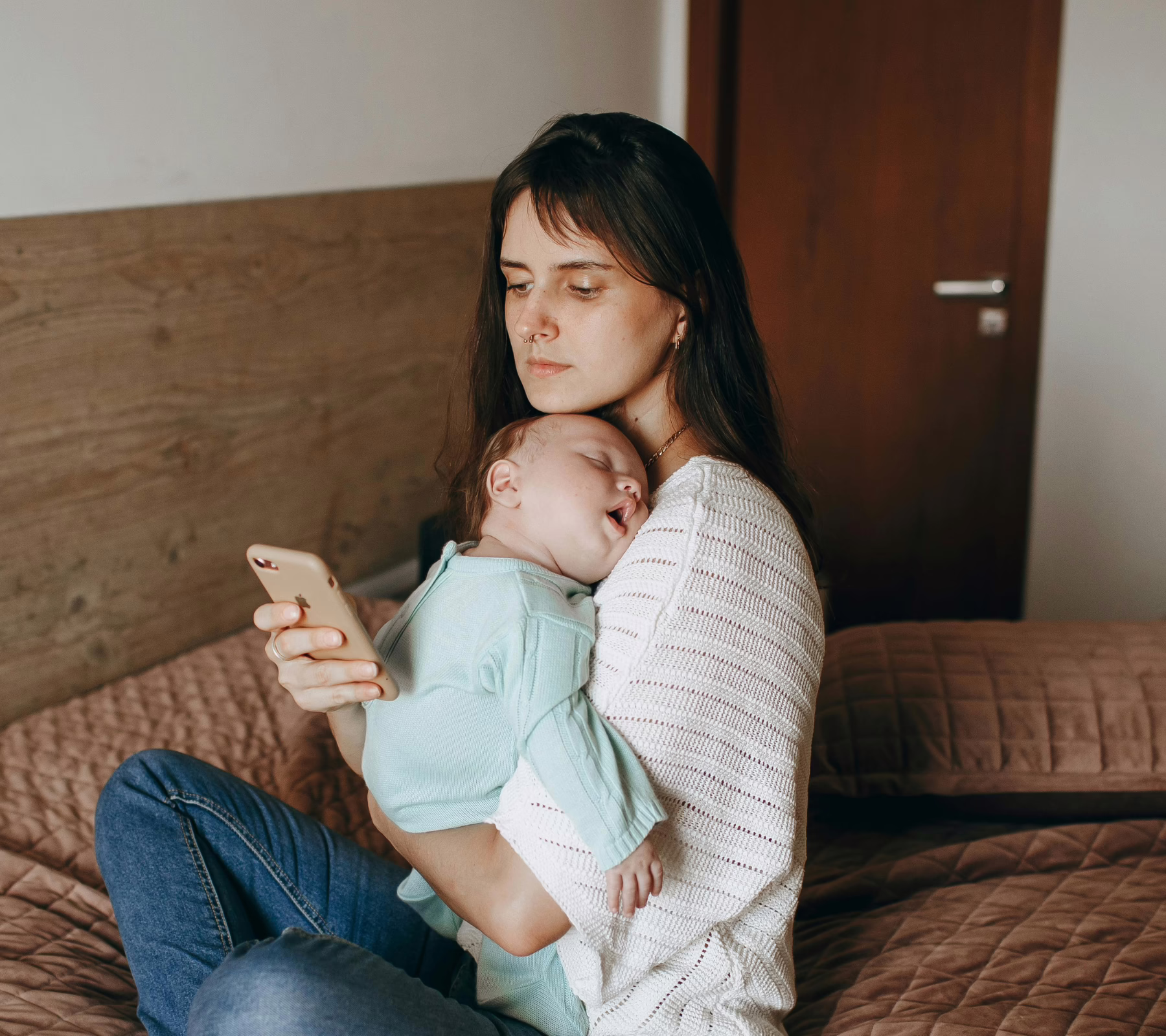 A young woman sits on a bed holding a sleeping baby against her chest while looking at her phone. The baby, dressed in a light blue outfit, rests peacefully on her shoulder.