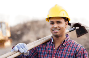 A construction worker wearing a yellow hard hat and gloves smiles at the camera while holding a shovel over his shoulder. He is dressed in a red plaid shirt, standing outdoors at a worksite.