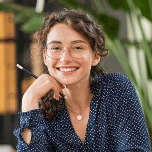 A young woman with curly brown hair and glasses smiles warmly while holding a pencil near her chin. She wears a navy blouse with white polka dots and a gold necklace, sitting in a bright indoor setting with blurred plants in the background.
