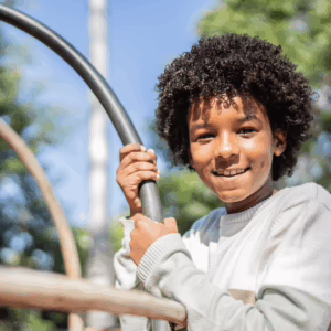 A smiling child with curly hair holds onto metal bars at a playground, with trees and blue sky in the background.