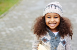 A young Black child with curly hair stands on a paved path, wearing a striped knit hat and a patterned sweater, holding a yellow book or folder against her arm.