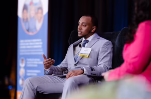 A speaker in a gray suit labeled “Jamal” addresses an audience from a stage, seated beside another panelist, with a Family Law CASA banner in the background.