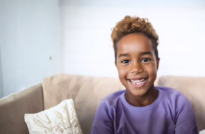 A young child with short curly hair sits on a beige couch indoors, wearing a light purple shirt against a softly lit neutral background.