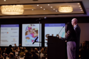 Family Law CASA’s Board President and Vice President speak at a podium during the luncheon, with sponsor logos displayed on screens behind them.