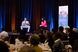 Audience members sit at round tables watching two speakers on stage, with blue and orange curtains and a Family Law CASA banner displayed beside them.